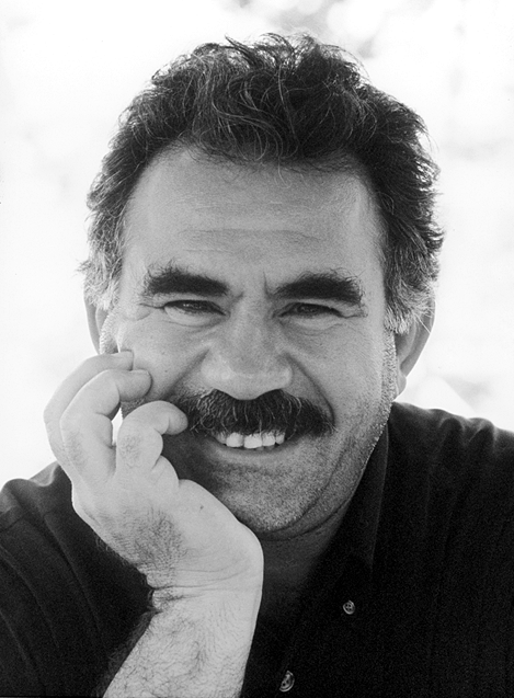 Black-and-white portrait of Abdullah Öcalan smiling, with a thick moustache and his cheek resting against one hand