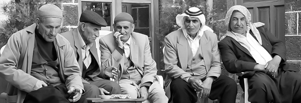 Five elderly dengbêj sit together outside a stone building in Diyarbakır in a black-and-white photograph