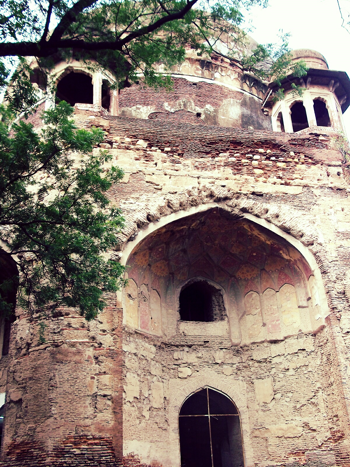 Weathered brick façade of Ali Mardan Khan’s tomb, with recessed arches, small rooftop pavilions and tree branches across the upper view