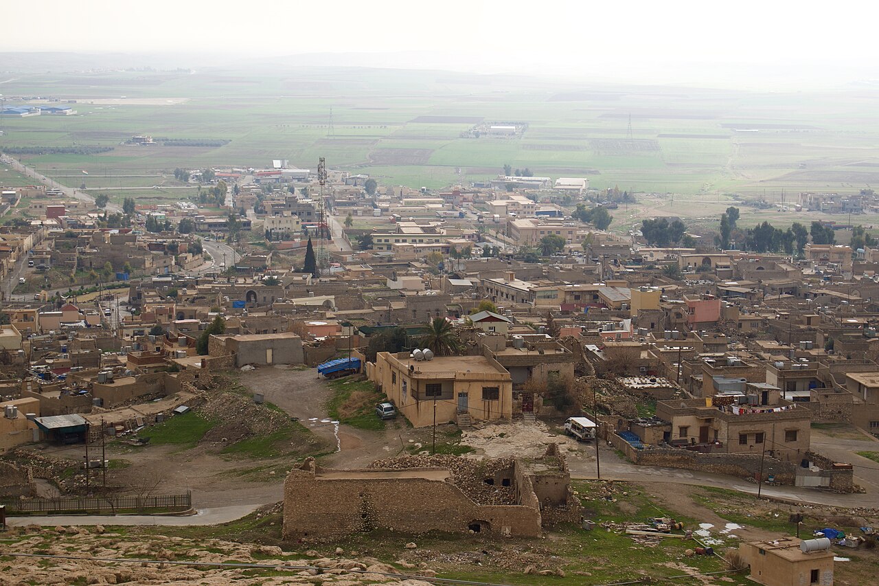 Stone and flat-roofed houses of Alqosh seen from above, with green agricultural fields across the plain beyond