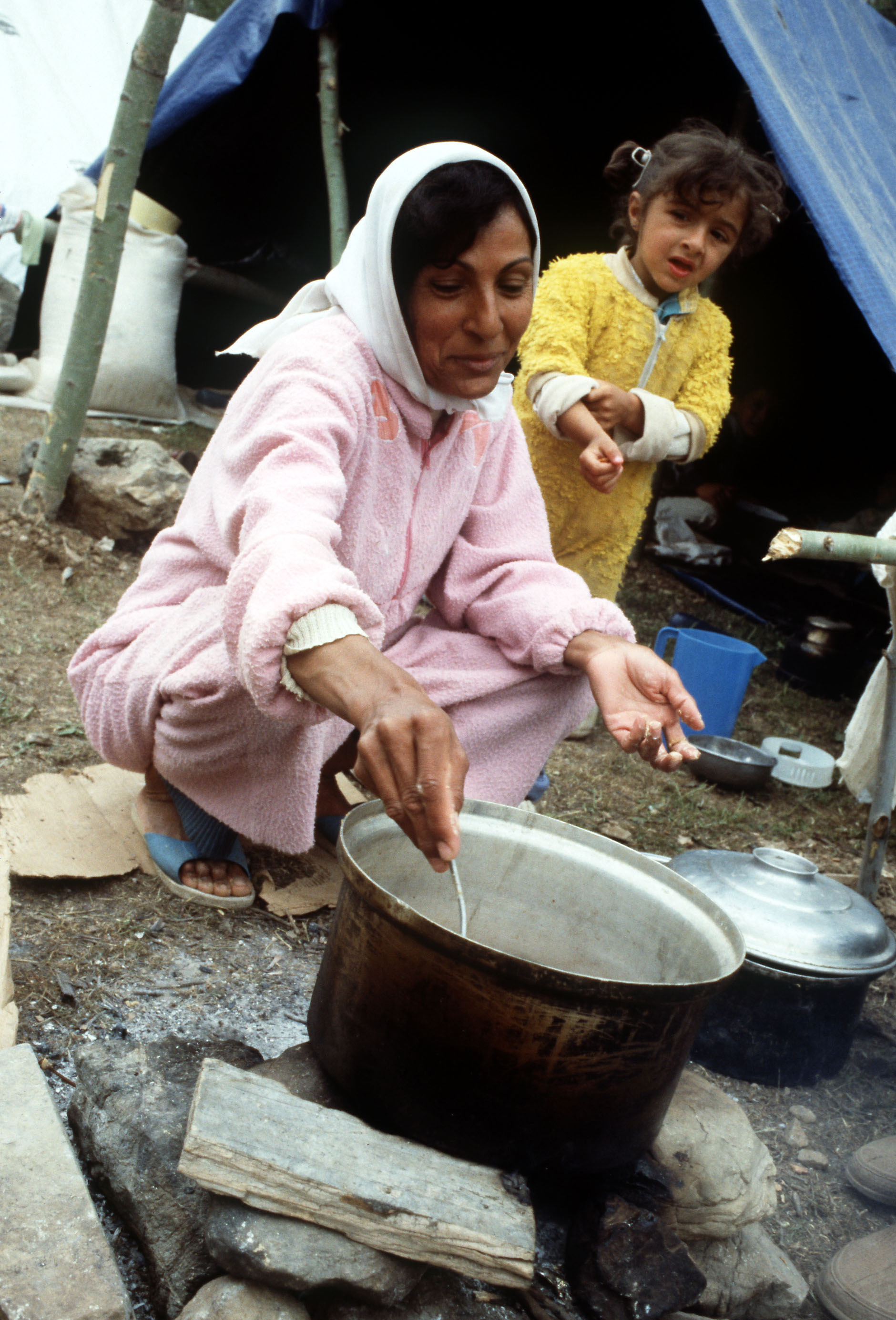 A woman in a pink cardigan stirs a cooking pot outside a blue tent while a girl in yellow stands beside her