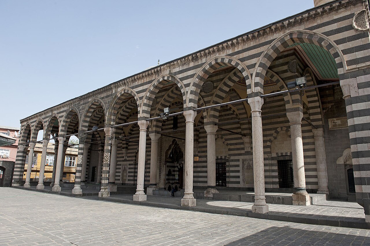 Pointed arches and columns of Behram Pasha Mosque’s portico, with alternating dark and pale stone bands and the entrance behind