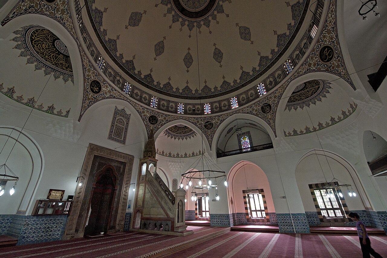 Interior beneath the decorated dome, with arched galleries, coloured windows, blue lower-wall tiles, a mihrab and a stone minbar