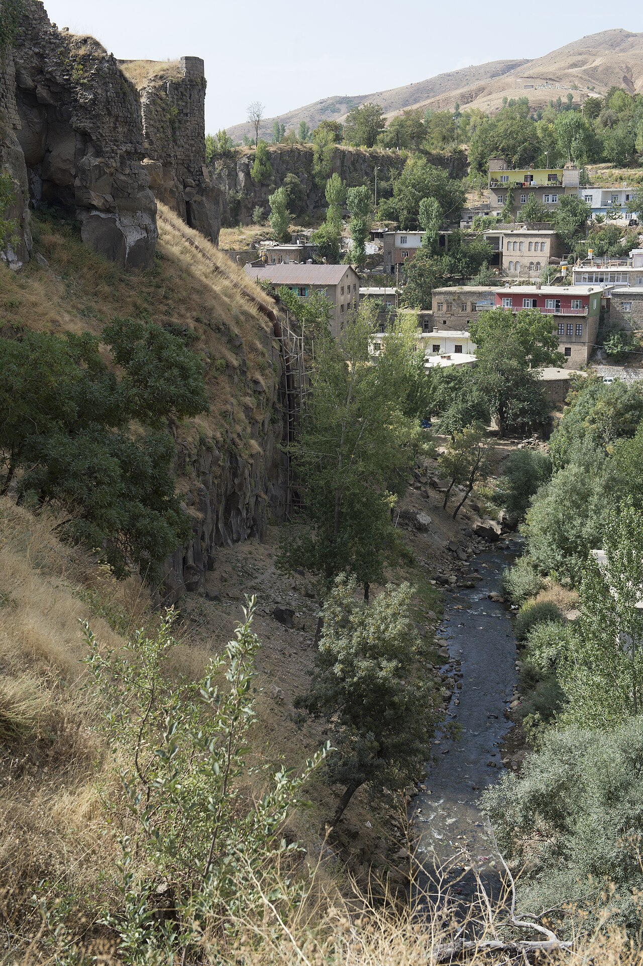 Murailles du château de Bitlis sur une paroi rocheuse au-dessus d’un ruisseau, avec arbres, maisons et collines sèches en face