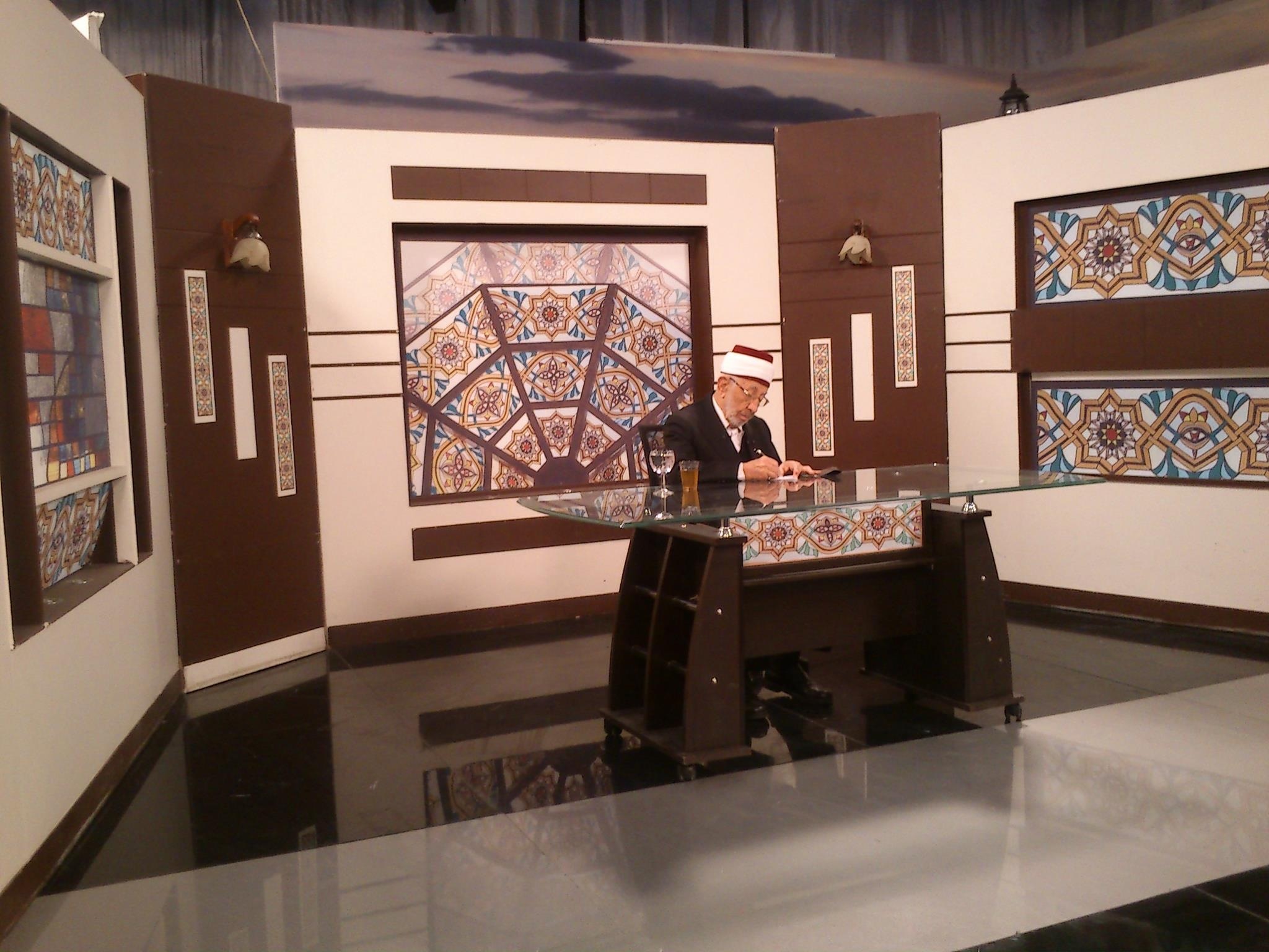 Muhammad Said Ramadan al-Bouti, wearing glasses and a white cap with a red top, writes at a glass desk in a studio decorated with geometric patterns