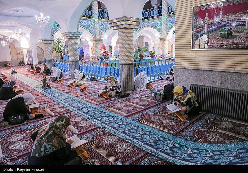 Women reading from books on wooden stands in a carpeted hall with spiral stone columns, tiled arches and a blue partition