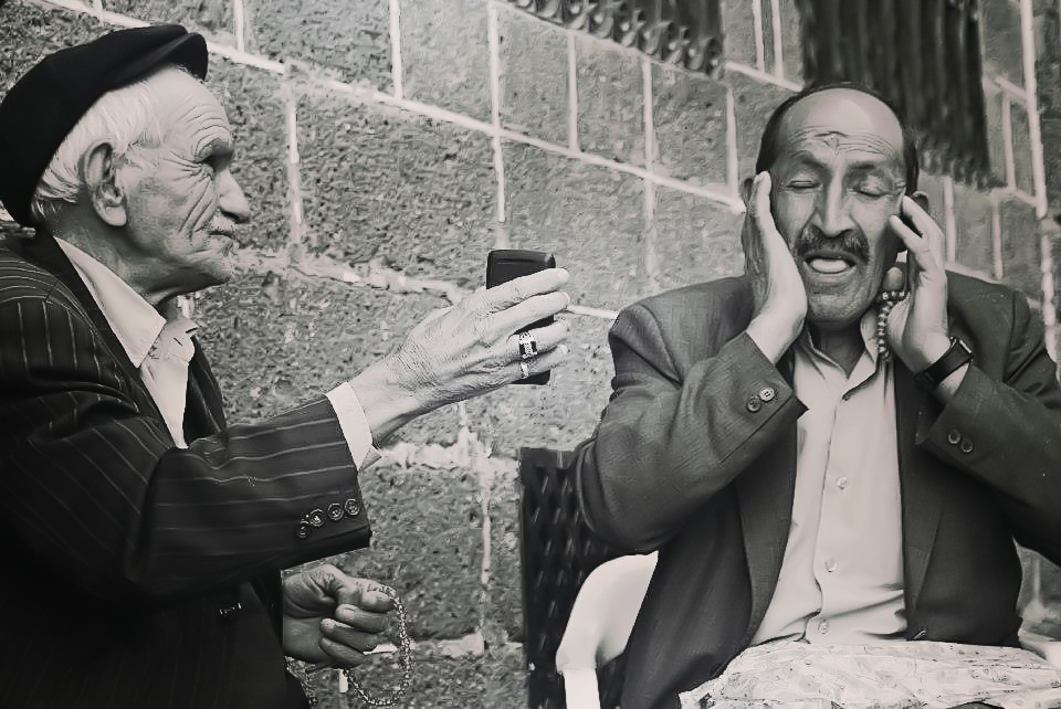 Two seated men beside a stone wall in Diyarbakir, one singing with his hands beside his face while the other holds a phone and prayer beads