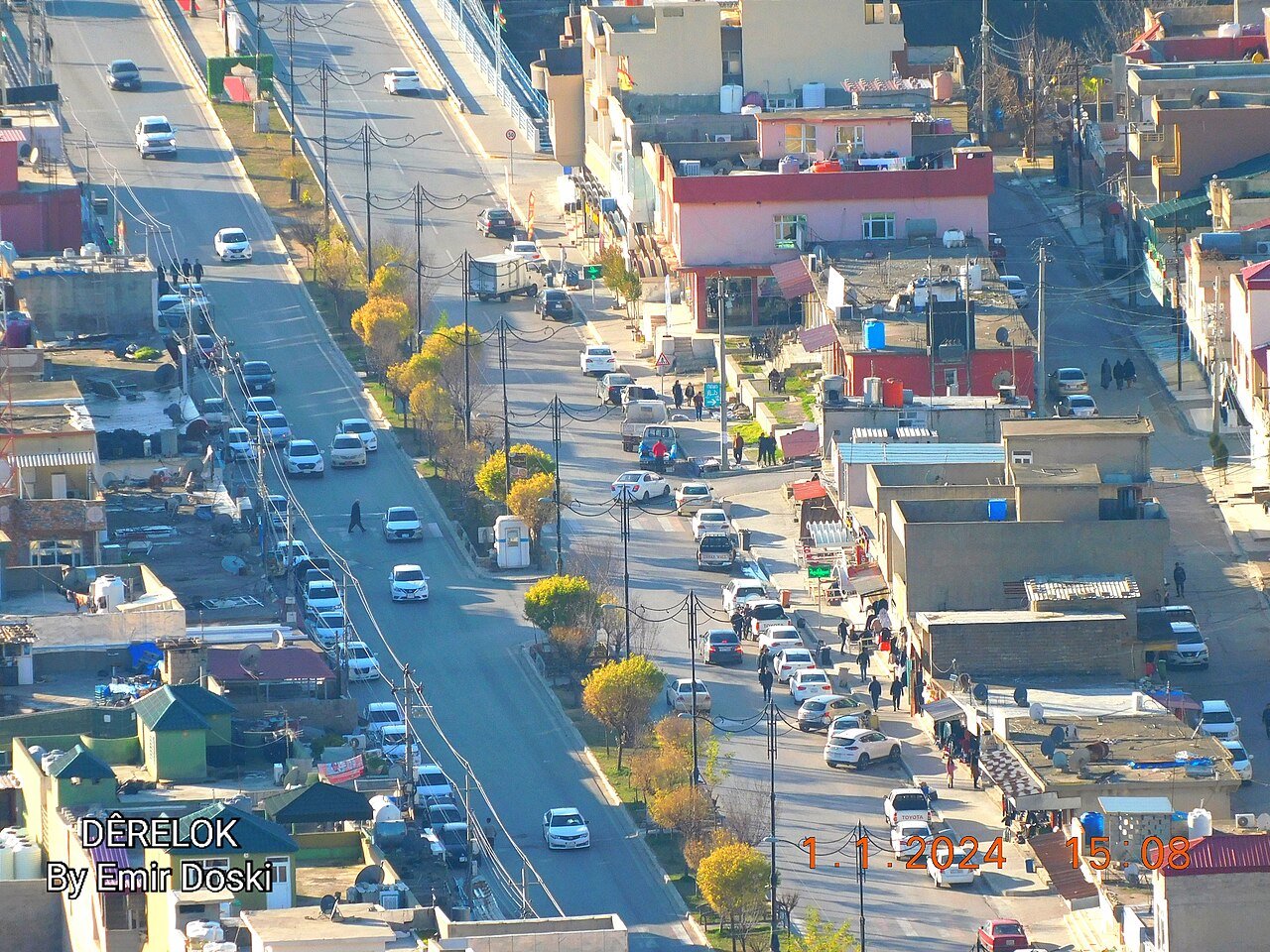 Elevated view along a divided street in Deralok with cars, pedestrians, utility wires and closely spaced flat-roofed buildings