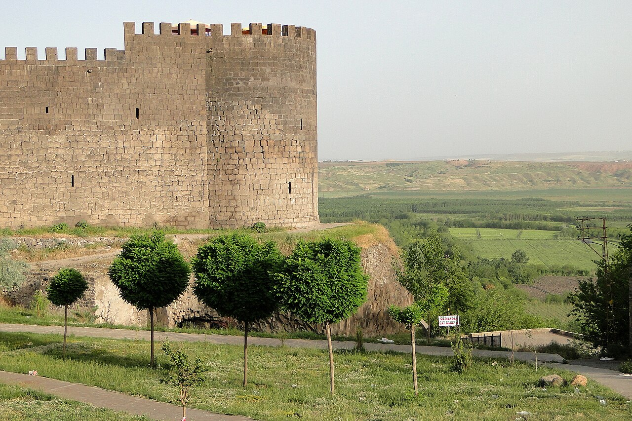 Round crenellated tower of Diyarbakır’s stone walls above clipped trees, with green cultivated land stretching into the distance