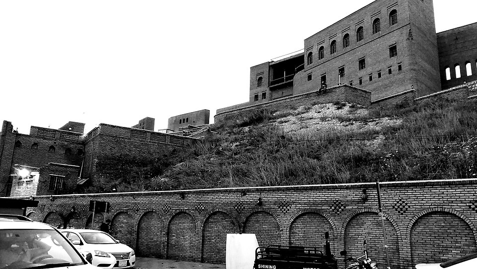 Schwarzweißfoto der Zitadellengebäude von Erbil über einem begrünten Hügel, darunter eine Ziegelmauer und parkende Autos