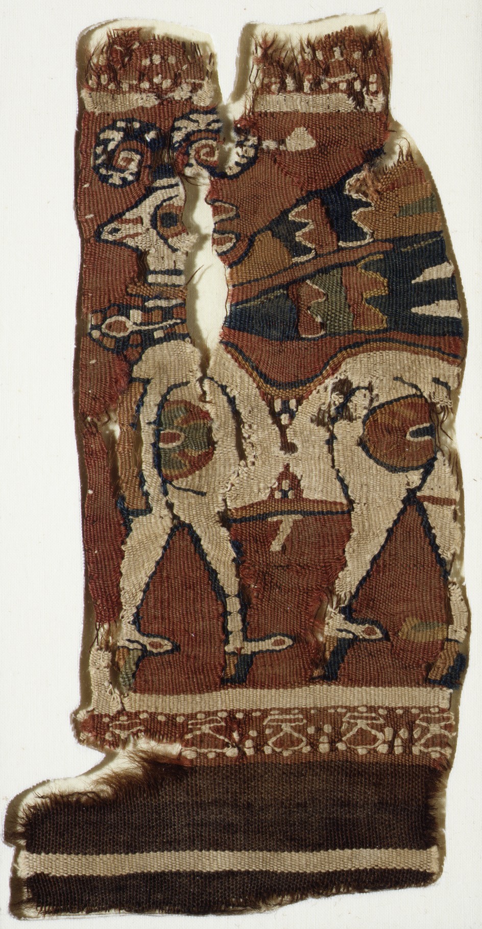 Fragment of red-brown woven cloth with a pale ram facing left, curled horns, a patterned neckband and flowing ribbons, divided by losses in the fabric