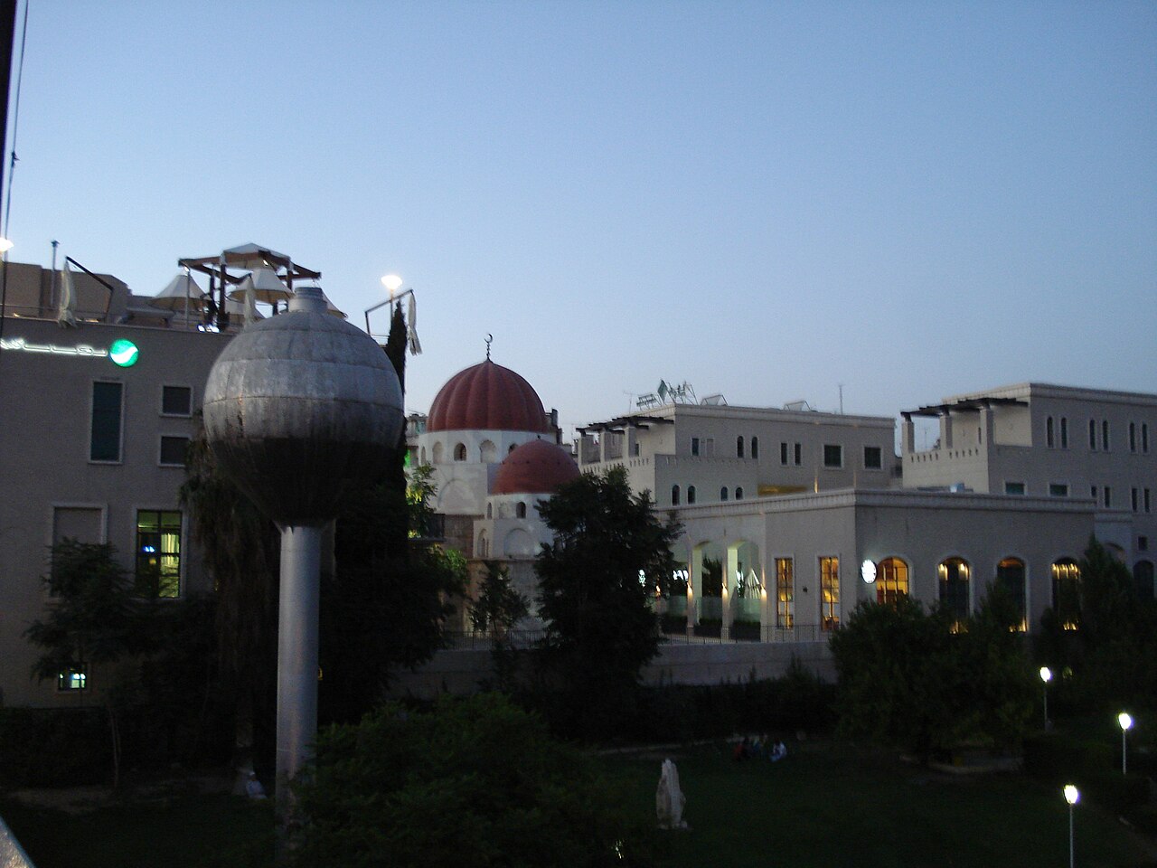 Coupoles rouges du complexe de Farrukh Shah à Damas parmi des bâtiments modernes clairs, avec arbres et lampadaire sphérique