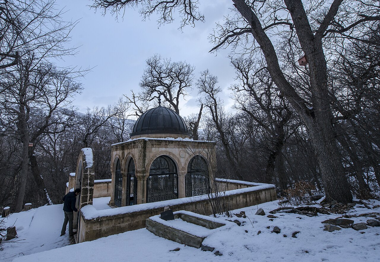 کوشک سنگی گنبددار با طاقها و دروازههای آهنی میان درختان بیبرگ و برف در آرامگاه منسوب به فقی طیران در ورزوز
