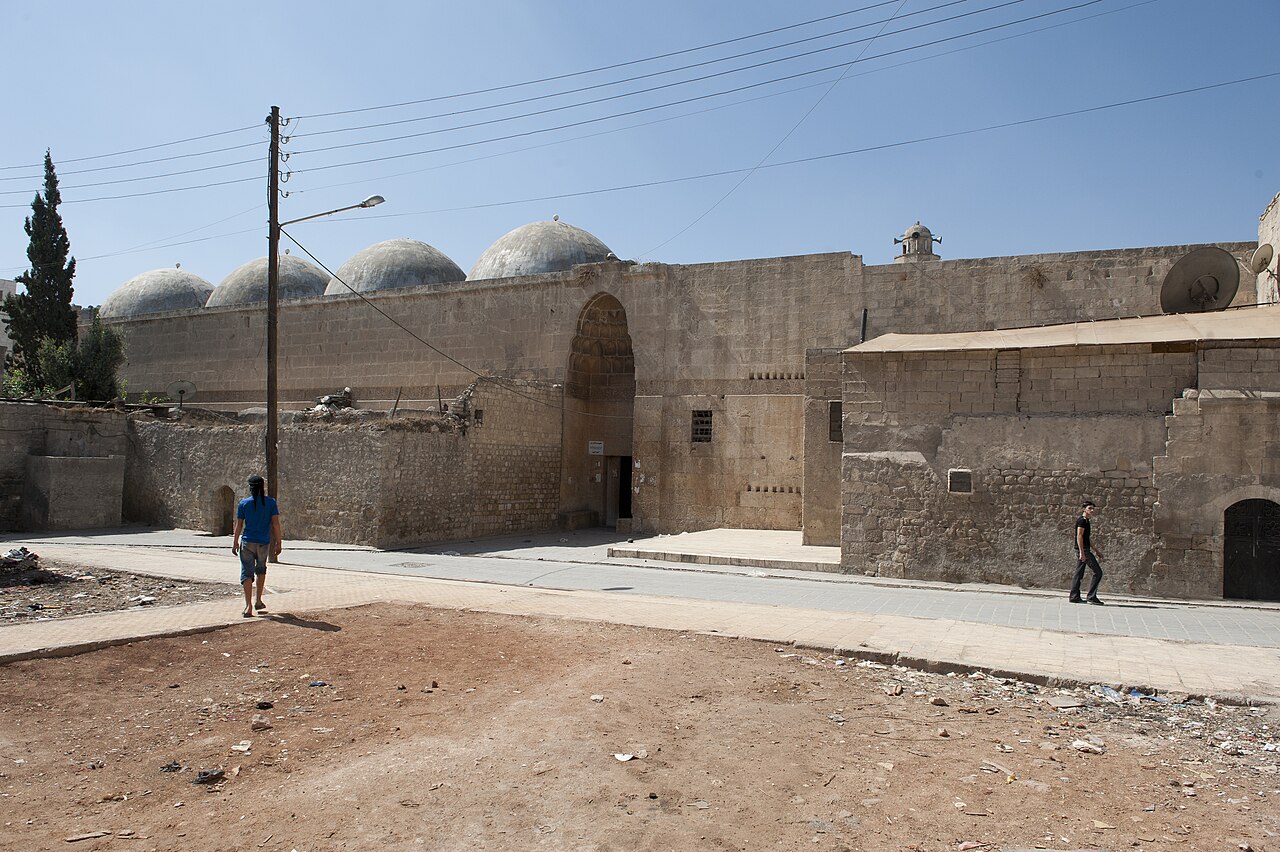 Buitenzijde van al-Firdaws in Aleppo met hoog muqarnasportaal in een stenen muur en vier zichtbare koepels