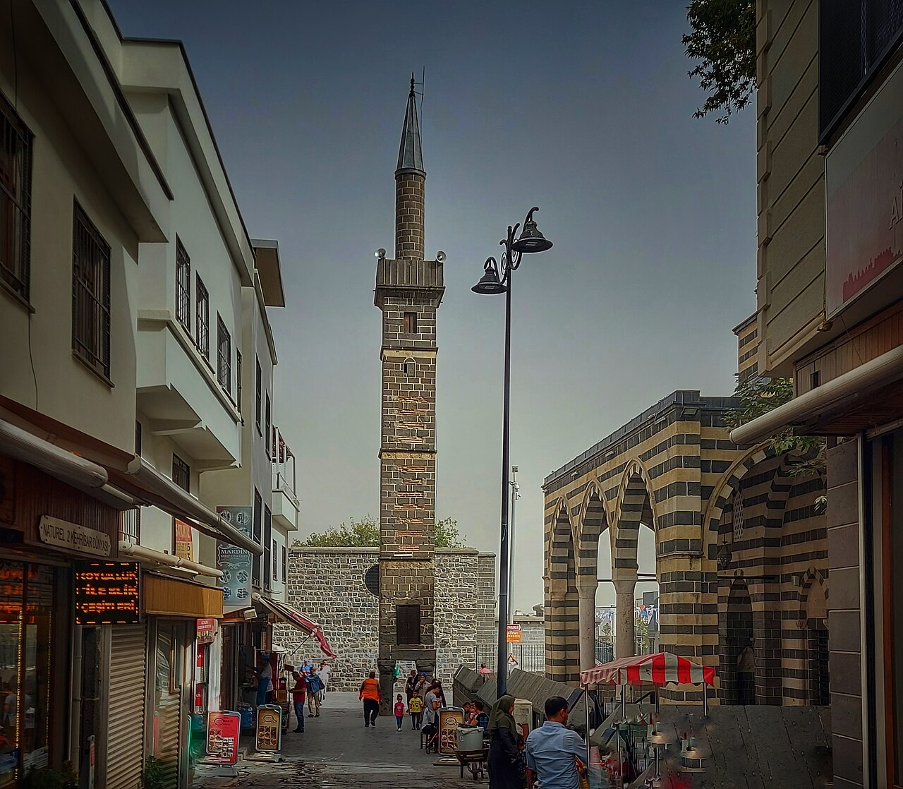 The Four-Legged Minaret rising above pedestrians in a narrow Diyarbakır street, with the striped stone arches of Şeyh Mutahhar Mosque on the right