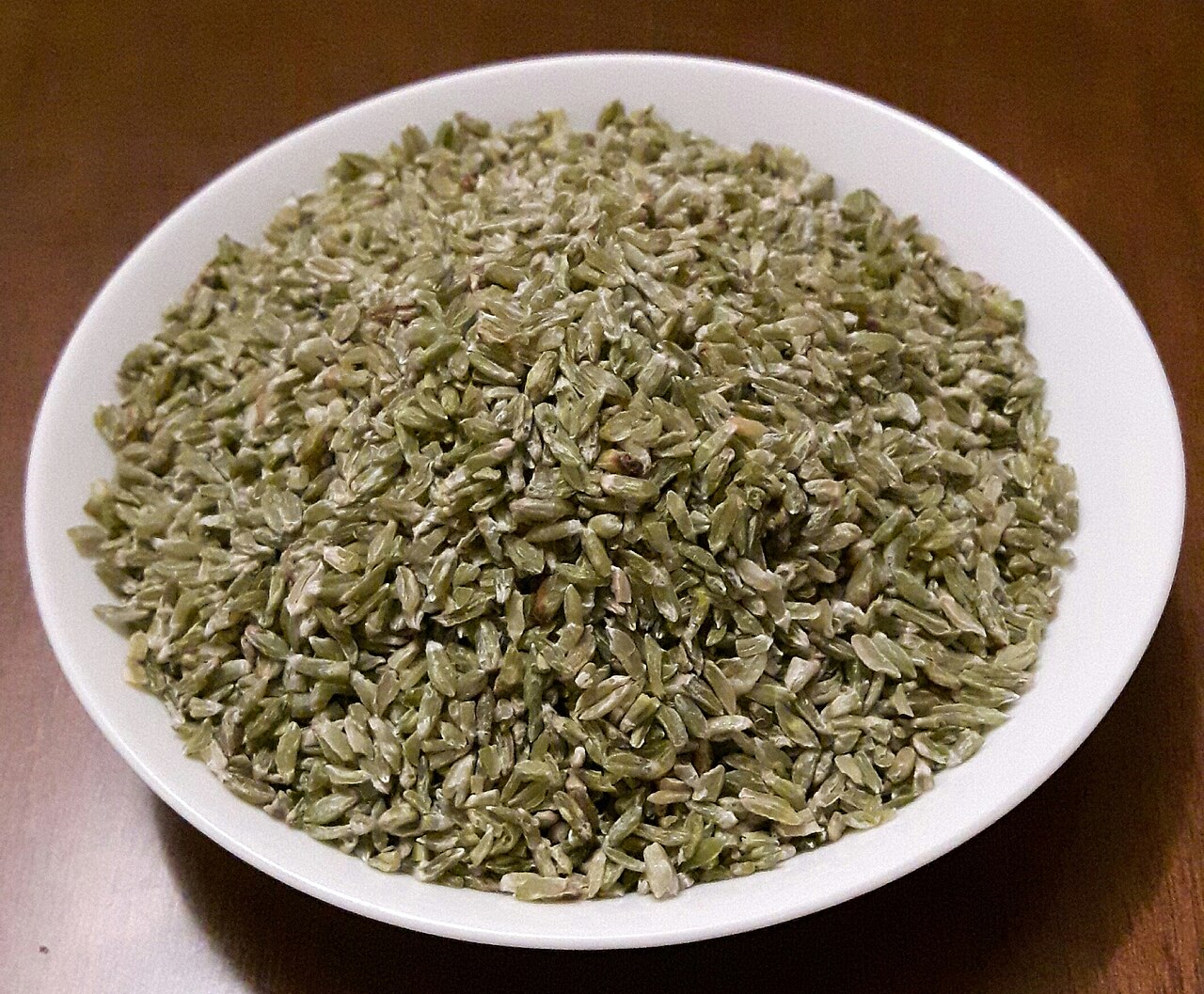 A white bowl filled with uncooked green-grey roasted wheat kernels on a brown wooden surface