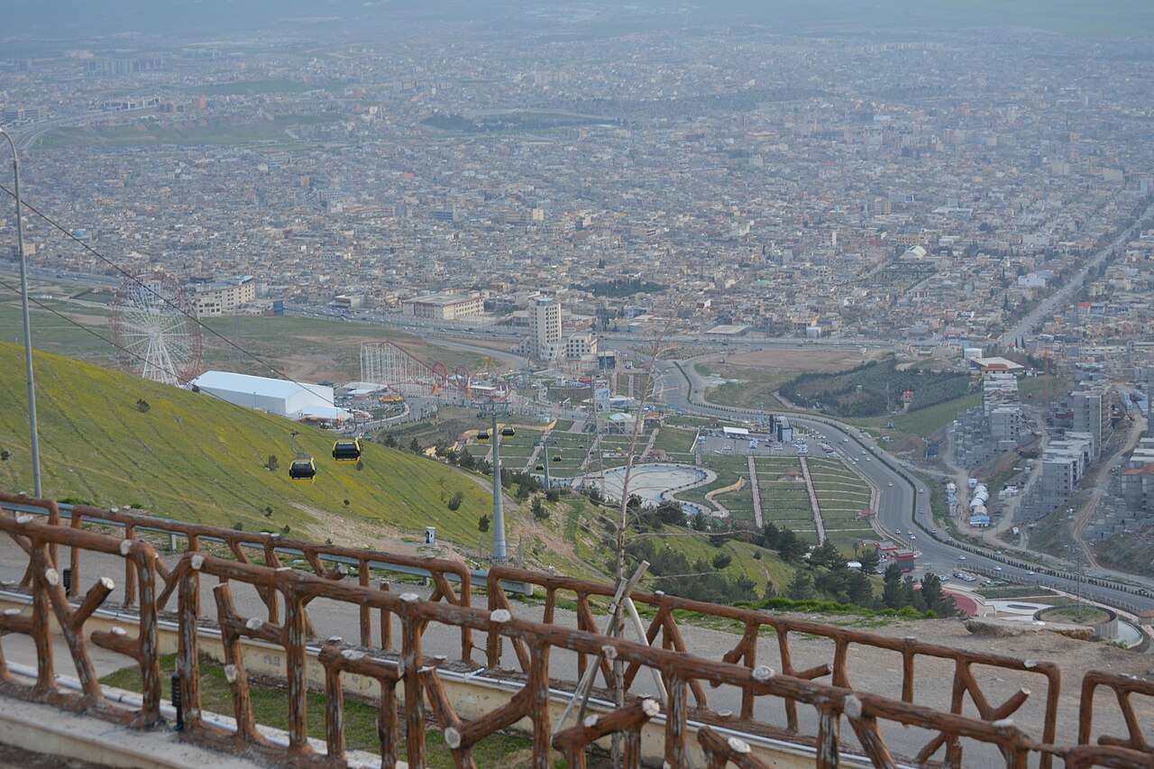 Vista desde una ladera verde de Goizha con telecabinas hacia la noria y atracciones de Chavi Land y los edificios densos de Sulaymaniyah