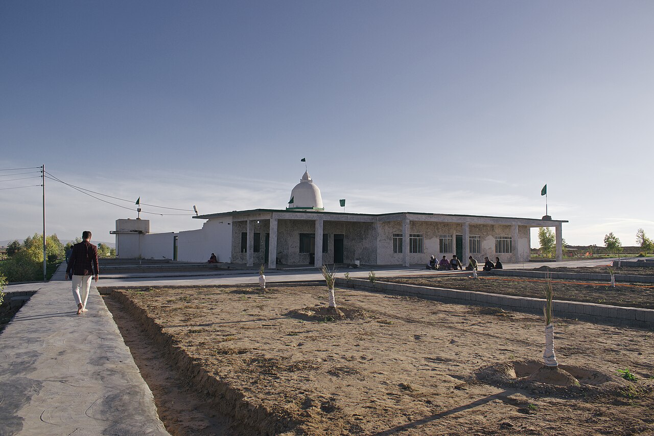 The low Shah Hayas Kakai shrine complex at Wardik with a small dome, green flags, visitors, paved paths and young trees