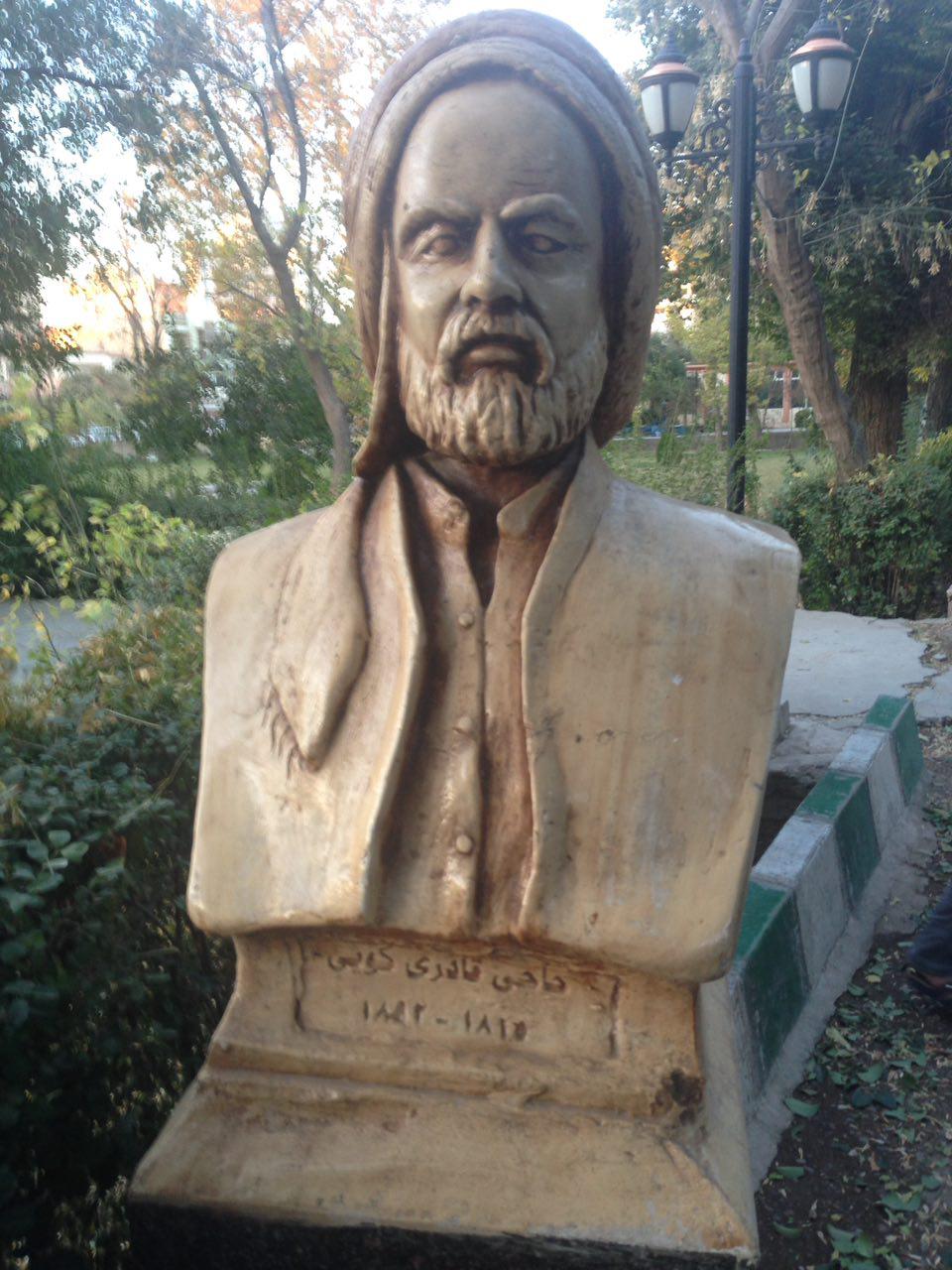 Memorial bust of Haji Qadir Koyi wearing a wrapped turban and buttoned jacket, on an inscribed plinth beside a garden path