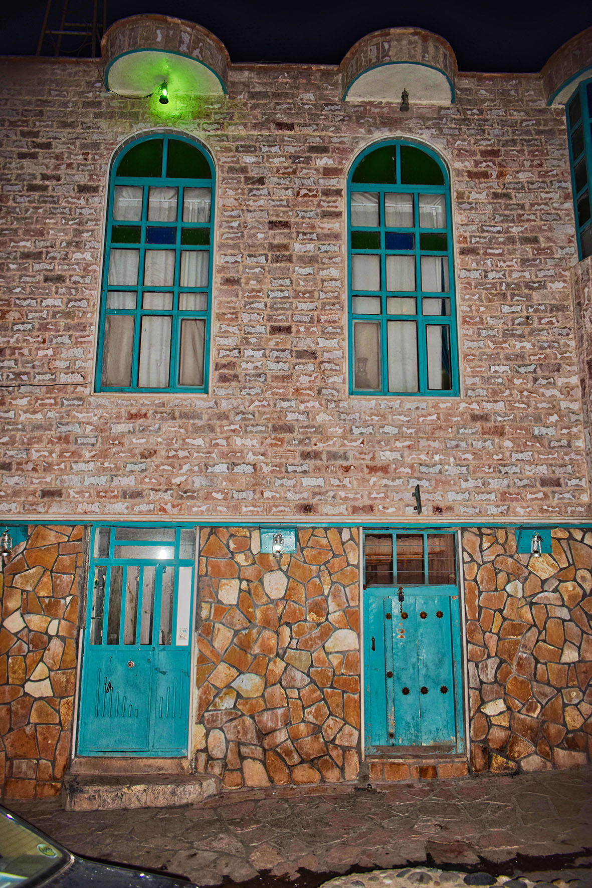 Stone and brick façade in Hajij with two tall turquoise arched windows, turquoise doors and a paved passage in front