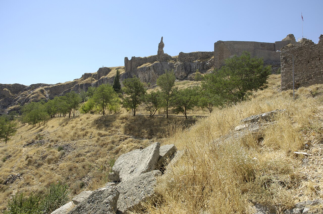 Harput Castle’s stone walls and ruined towers above a rocky slope with dry yellow grass, scattered green trees and fallen stone blocks