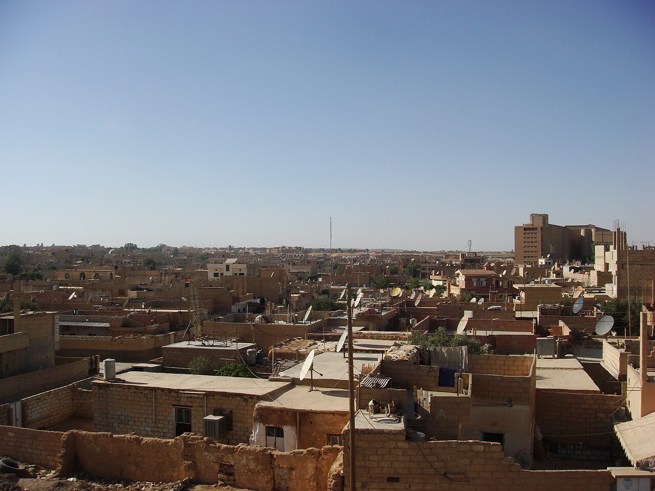 Flat-roofed houses, rooftop satellite dishes and a taller building in Hasakah’s Tal Hajar quarter beneath a clear sky