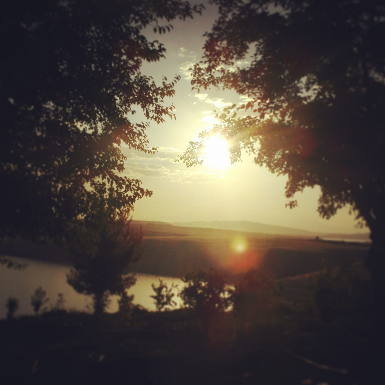 Low sun over a broad stretch of water and distant hills, framed by dark trees in a photograph labelled Hilvan