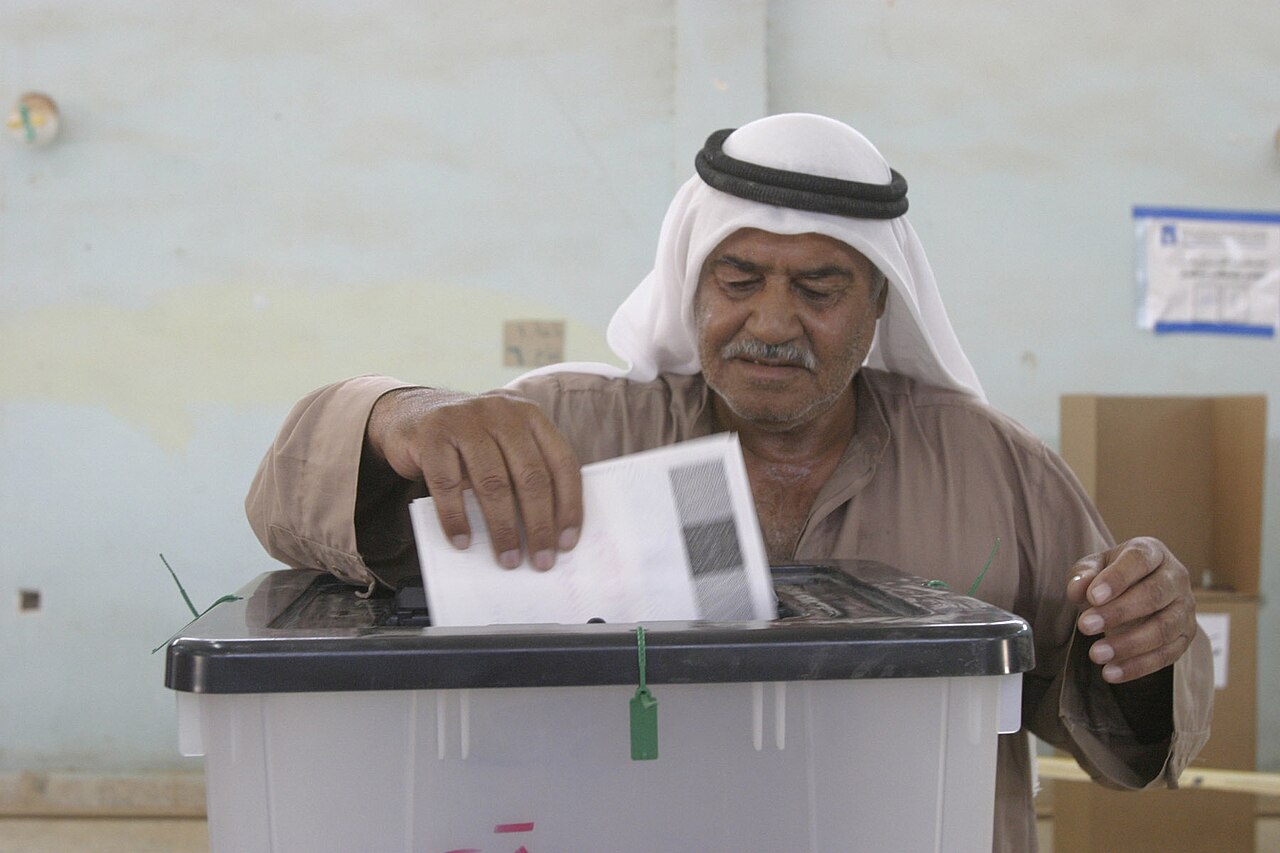 A man in a white headcloth and brown robe inserts a paper ballot into a sealed ballot box at a polling station in Barwana