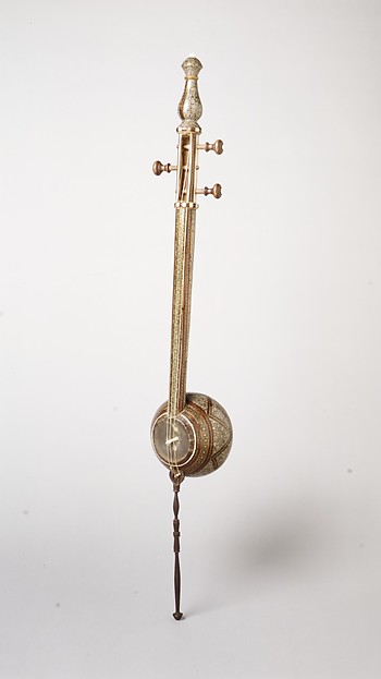 Historic kamancheh standing upright on a long metal spike, with a round inlaid resonator, slender neck and tuning pegs against a pale background
