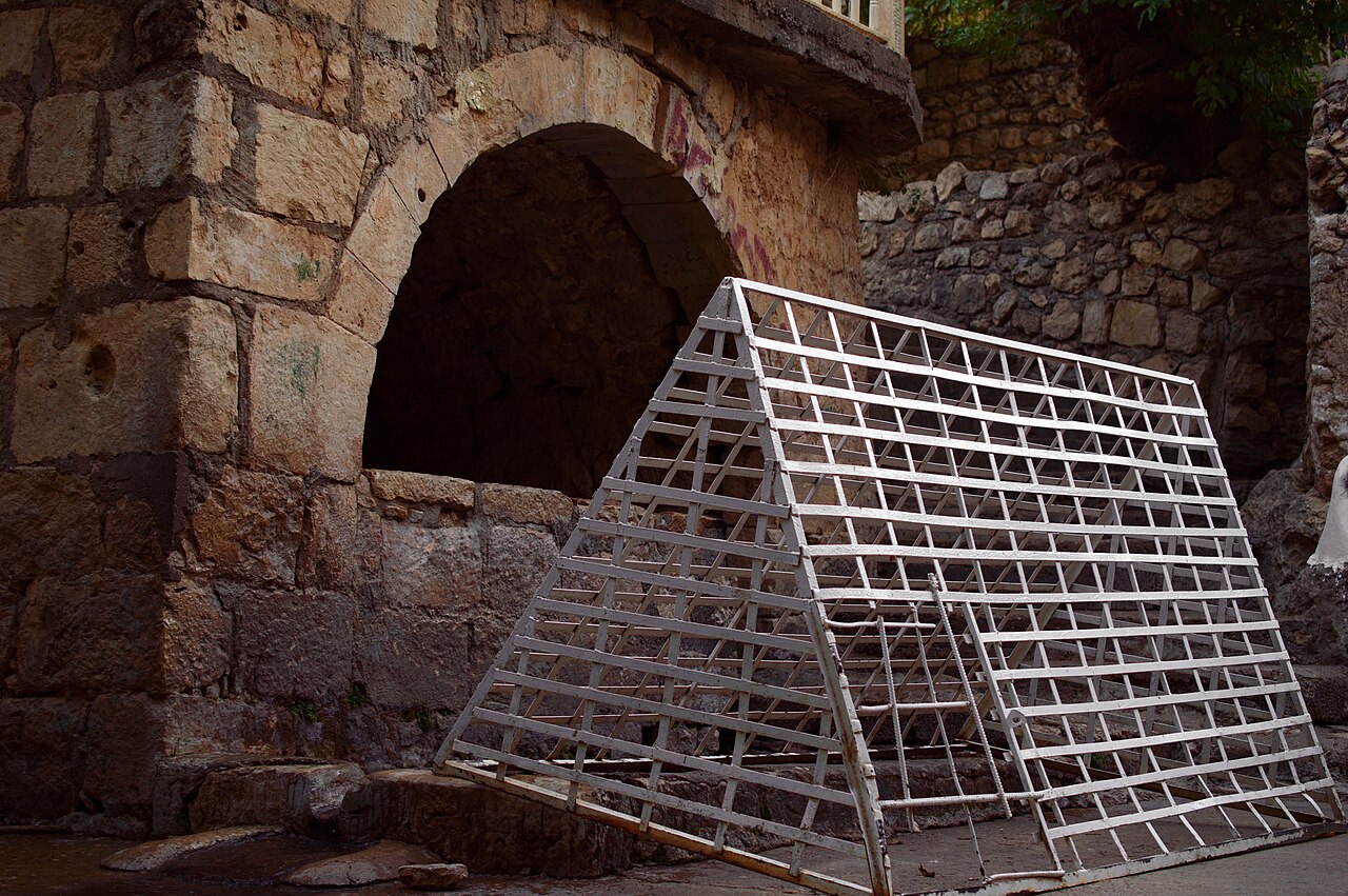 Steinbogen und niedriger Steinsims in Lalish, davor ein großes angelehntes weißes Metallgitter, Bäume über der Mauer