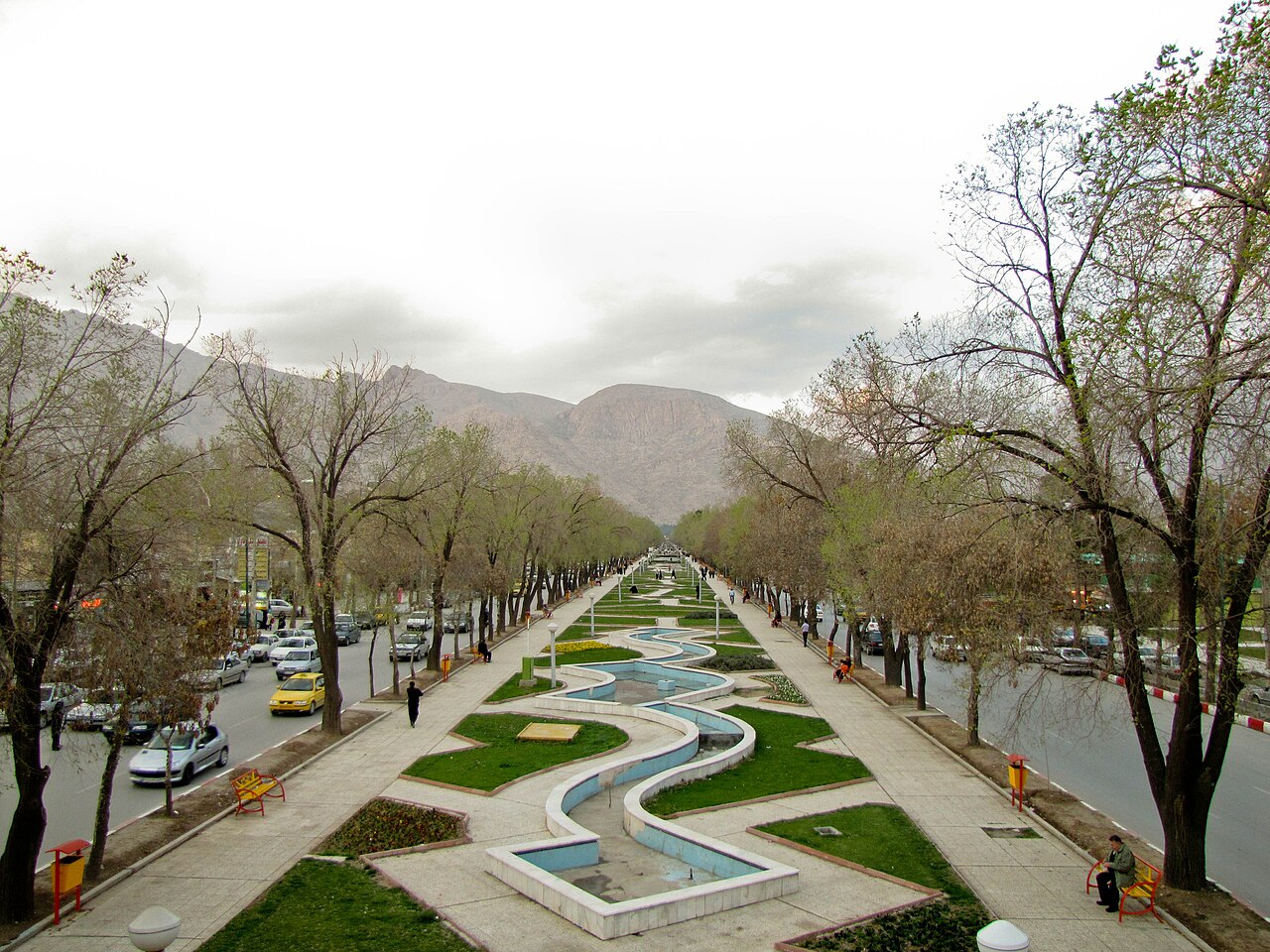 Trädkantad Shirudiboulevard i Kermanshah med slingrande vattenkanal, gående, bilar och avlägsna berg