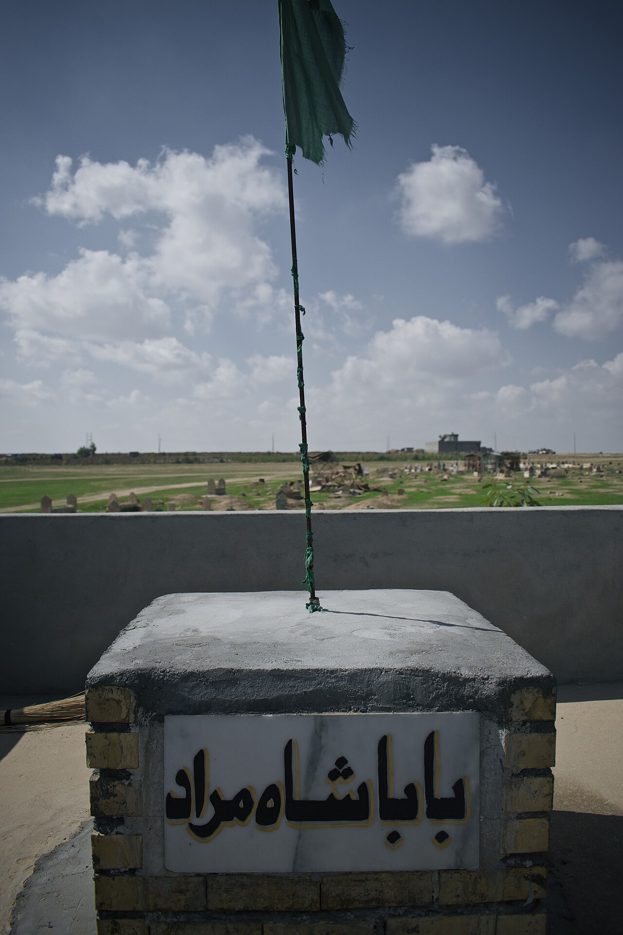 Petit marqueur maçonné inscrit portant une haute bannière verte au complexe kaka’i de Wardik, avec cimetière et champs verts au-delà d’un muret