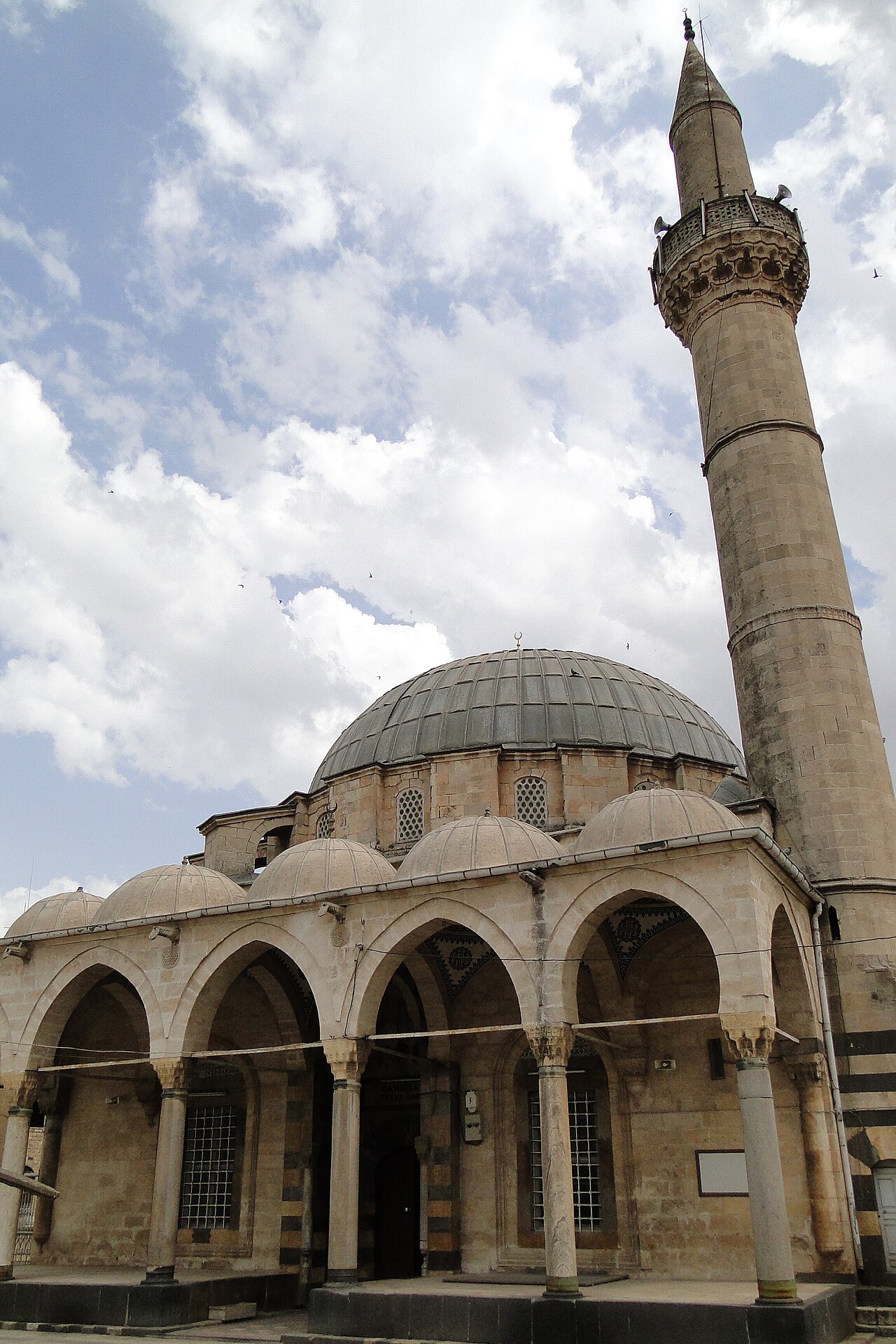 Kilis Tekke Camii; büyük kurşun kaplı kubbe, sivri kemerli revak üzerinde küçük kubbeler ve bulutlar önünde yüksek taş minare