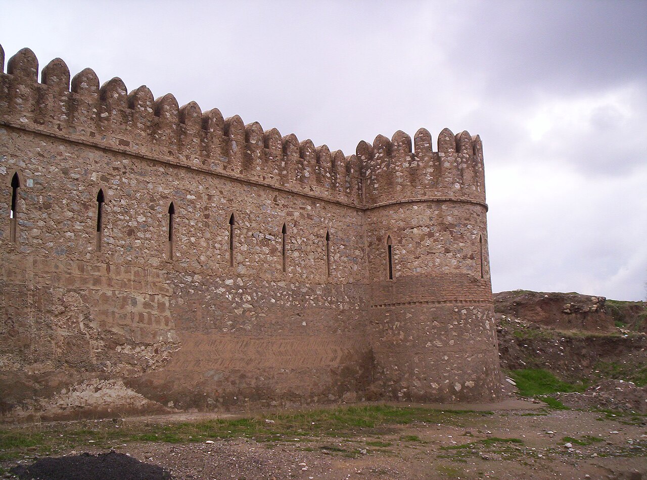 Weathered stone walls and rounded corner tower of Kirkuk Citadel with narrow openings and crenellations beneath a cloudy sky