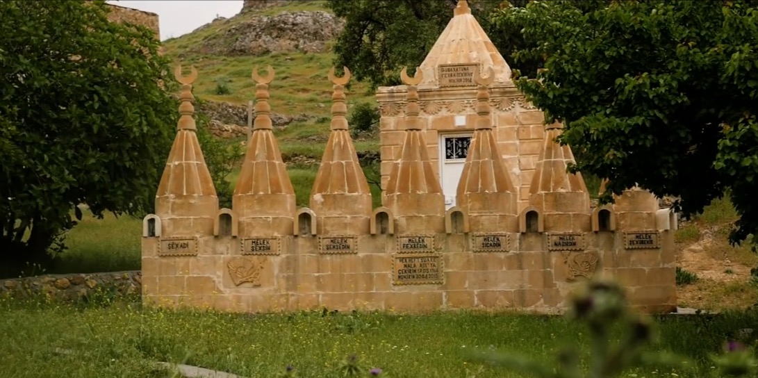 Adlandırılmış küçük konik kuleler ve hilal biçimli tepeler sırası bulunan taş Êzidî ziyaretgâhı, arkada daha büyük yapı ve iki yanda ağaçlar