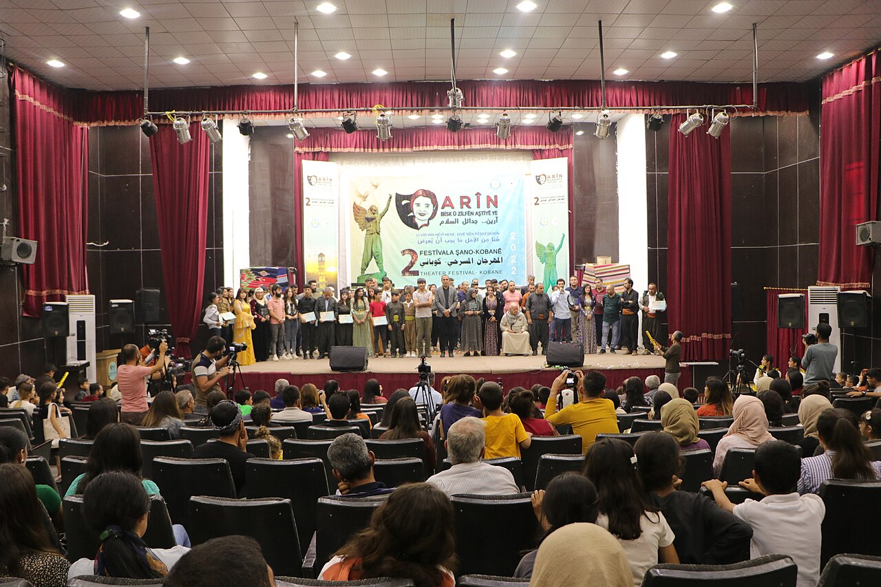 Audience facing a stage where participants stand beneath the Arîn theatre festival banner in Kobani in 2022