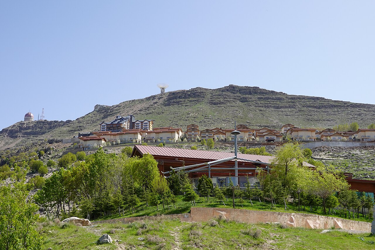 Korek resort buildings and green trees below a layered rocky ridge, with an observatory dome at left and a dish antenna higher on the slope