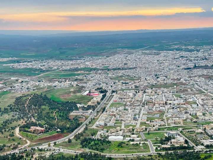 Elevated view over Koya showing dense pale buildings, roads, green plots and surrounding plains beneath a sunset sky