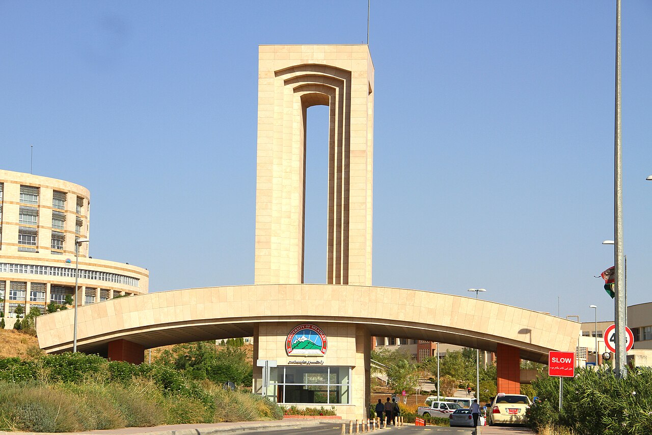 Heller steinerner Eingangsbogen und Turm der Universität Sulaimani, mit Menschen und Fahrzeugen am Tor