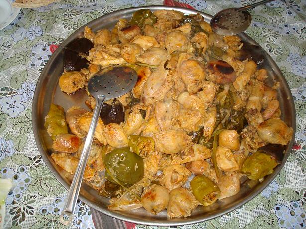 Gran bandeja metálica redonda con pimientos, cebollas, berenjenas y otras verduras rellenas y dos cucharas sobre un mantel estampado