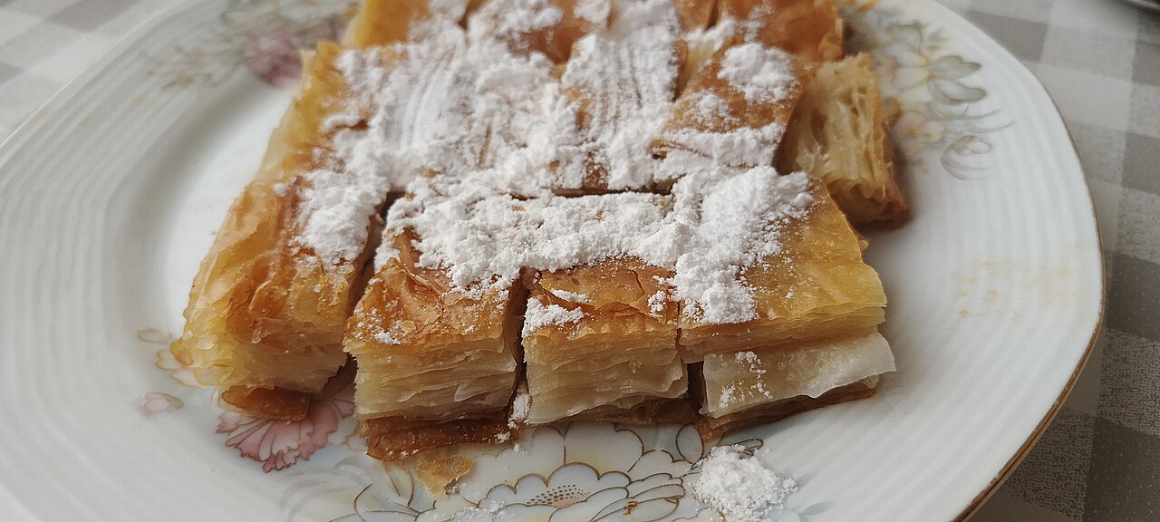 Rectangular pieces of golden layered Kürt böreği dusted with powdered sugar on a white floral plate, showing the thin unfilled pastry layers