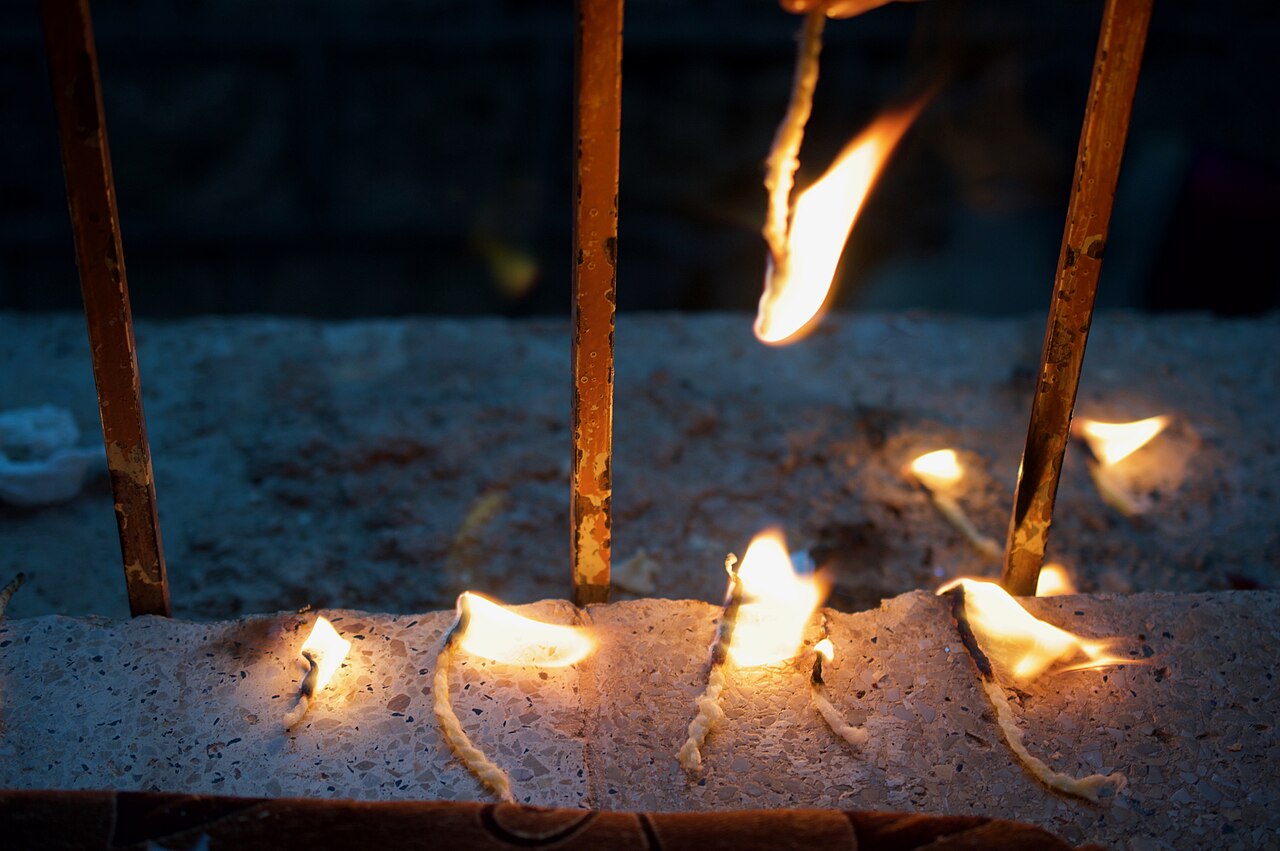 Kleine ölgetränkte Baumwolldochte brennen auf einer Steinkante in Lalish; dahinter eine größere Flamme zwischen senkrechten Metallstäben