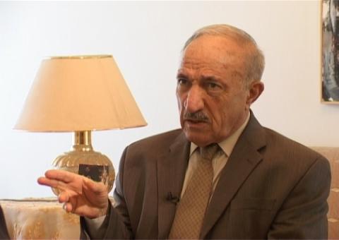 Mahmoud Othman assis en costume brun et cravate à motifs, parlant une main levée près d’une lampe