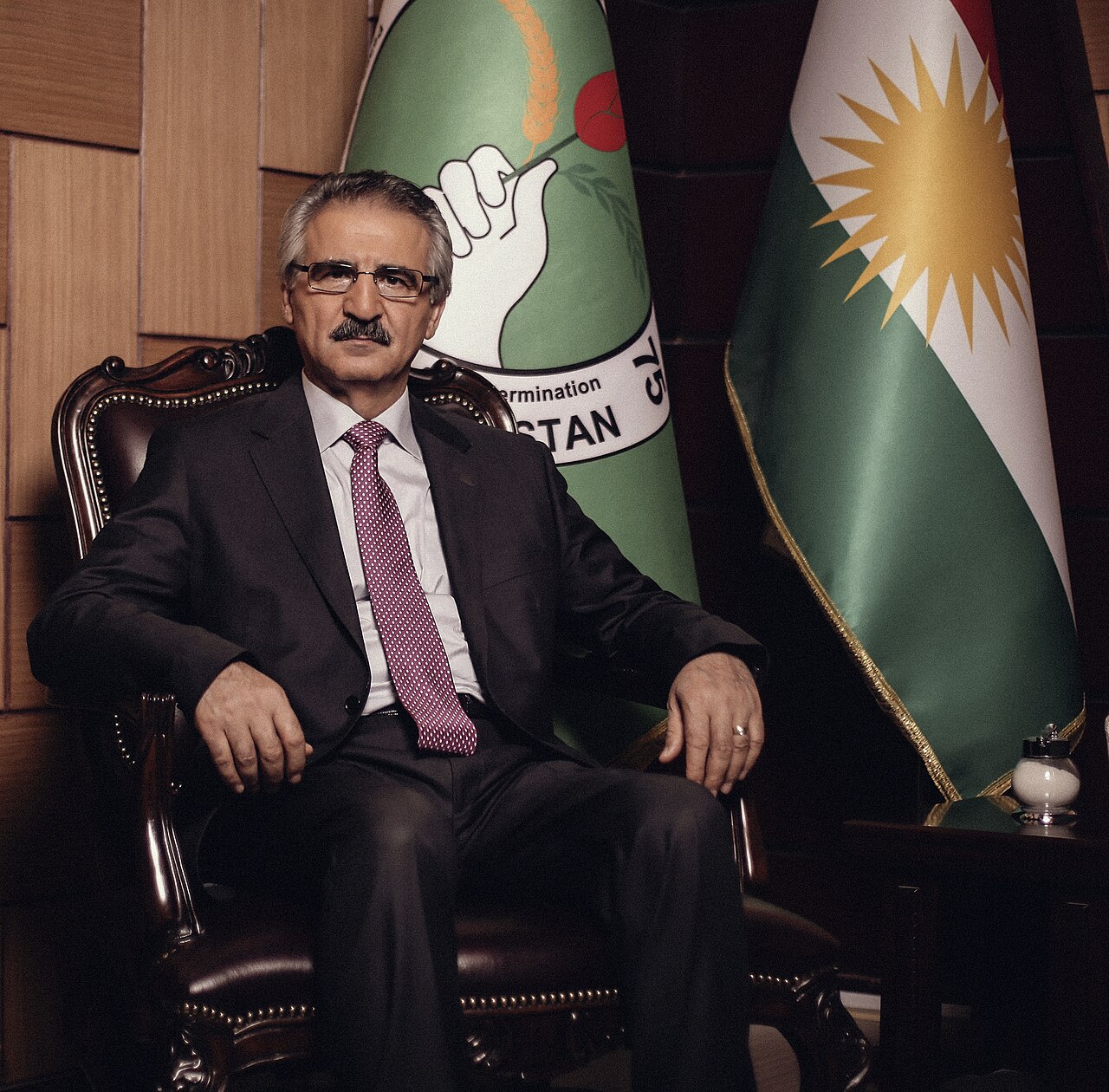 Mala Bakhtiyar mit Brille und dunklem Anzug im Sessel zwischen grünem PUK-Banner und Kurdistanflagge