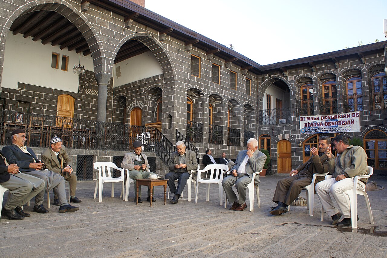 Men seated with tea in the courtyard of a dark stone house, beneath arches and a Mala Dengbêjan banner in Diyarbakır