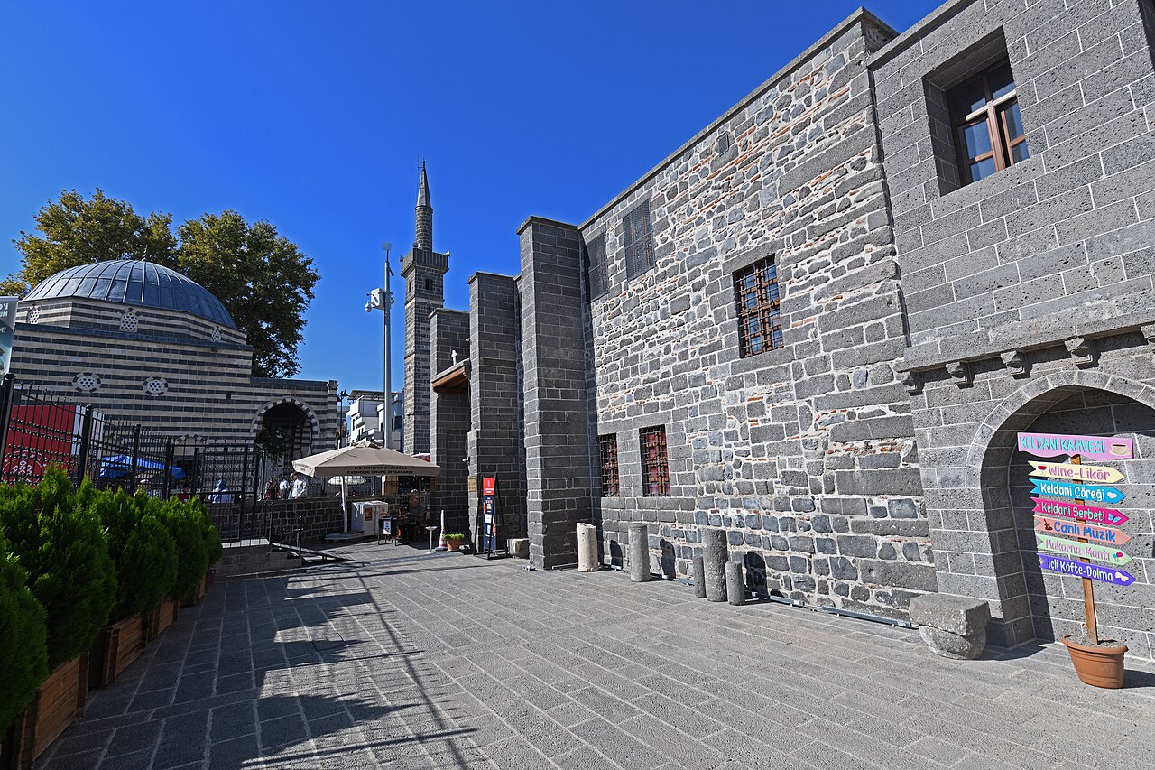 Dark basalt exterior of Mar Petyun Church with projecting buttresses and barred windows beside a paved lane, with a mosque dome and minaret nearby