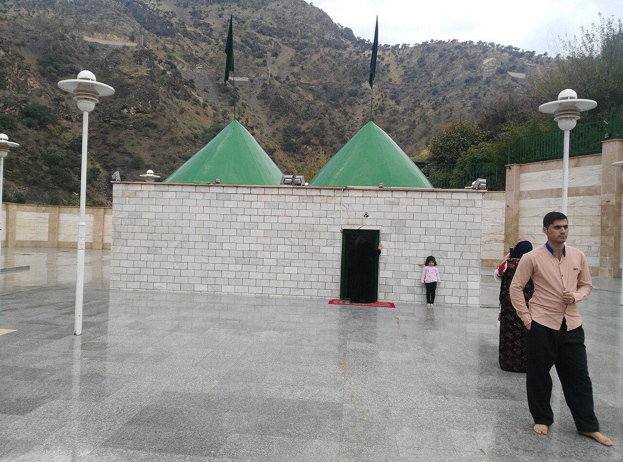 Two green conical roofs above the white-walled shrine of Sultan Sahak, with visitors in the courtyard and rocky mountains behind
