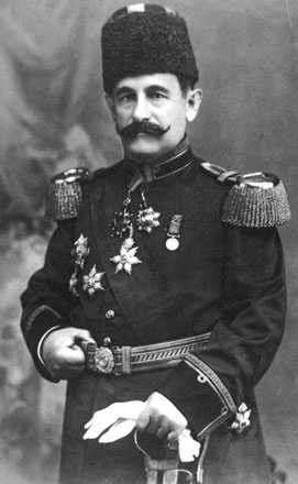 Portrait noir et blanc de Mustafa Yamulki, moustache, haute coiffe sombre, uniforme décoré, ceinture tressée et gants près d’une épée
