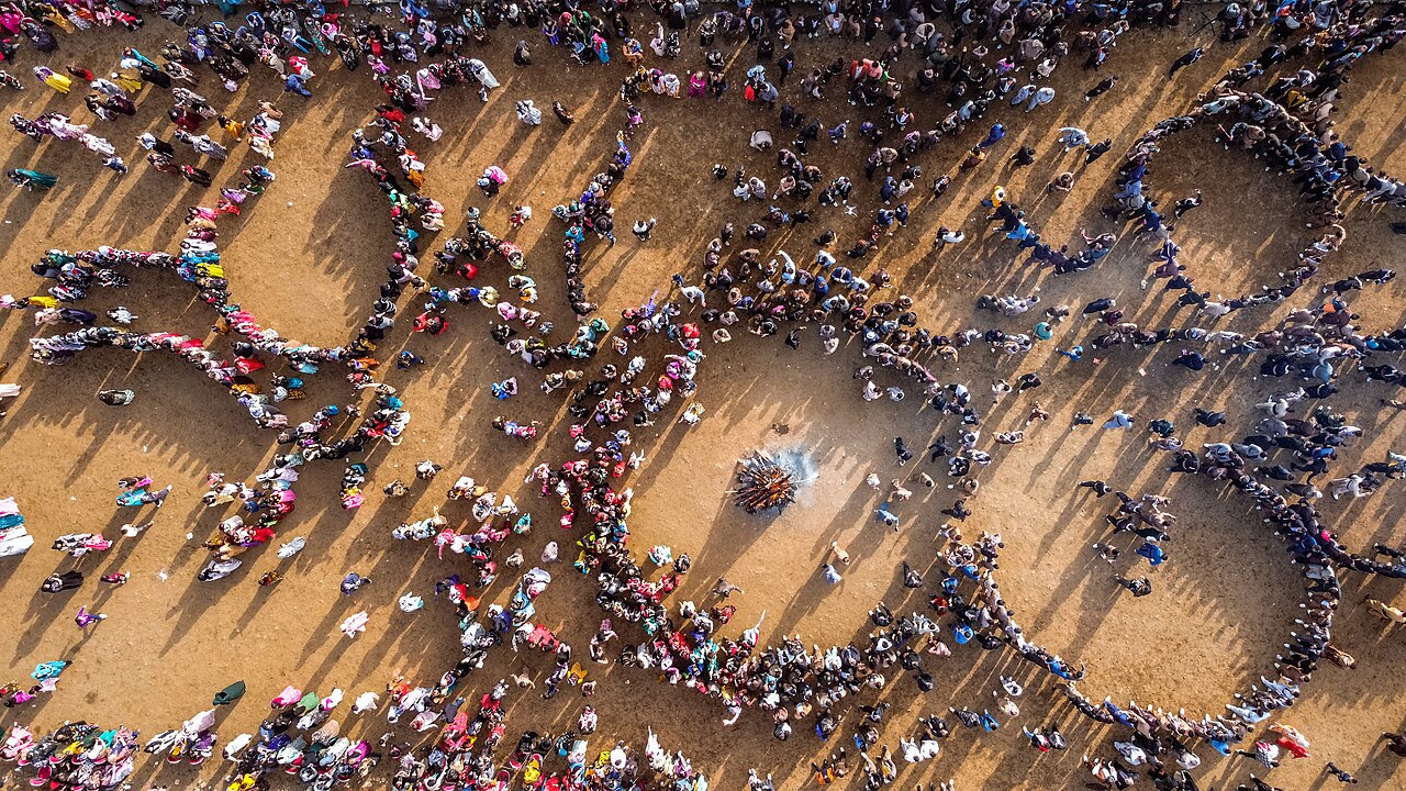 Aerial view of large circles and lines of people in colourful clothing dancing around a small smoking bonfire on open ground