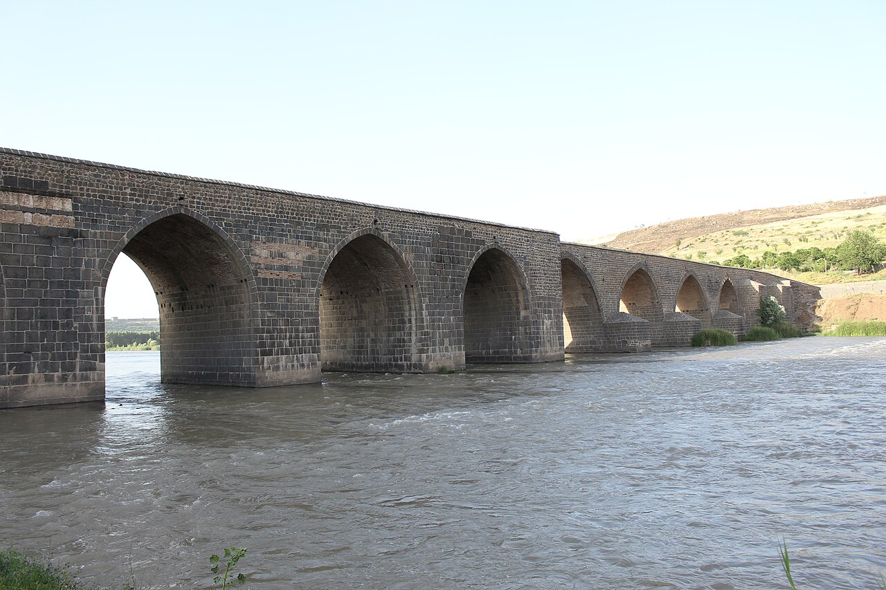 Dunkle Steinbögen der Dicle-Brücke über dem Tigris, Wasser im Vordergrund und sonniger Hang dahinter